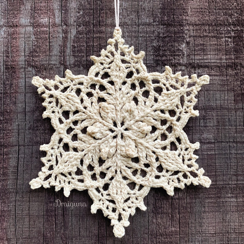 May include: A crocheted snowflake ornament in a cream color, hanging from a string against a brown wooden background.