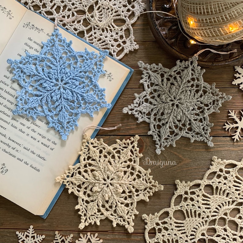 May include: Six crocheted snowflakes in various shades of blue, gray, and beige. The snowflakes are arranged on a wooden surface with a book and a spool of yarn.