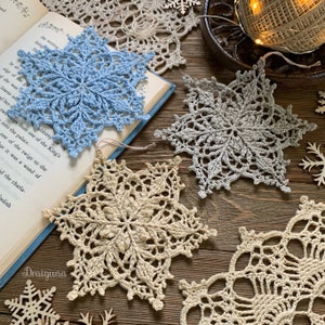 May include: Three crocheted snowflakes in different colors and patterns. The snowflakes are light blue, gray, and beige. They are arranged on a wooden surface with a book and other decorative items.