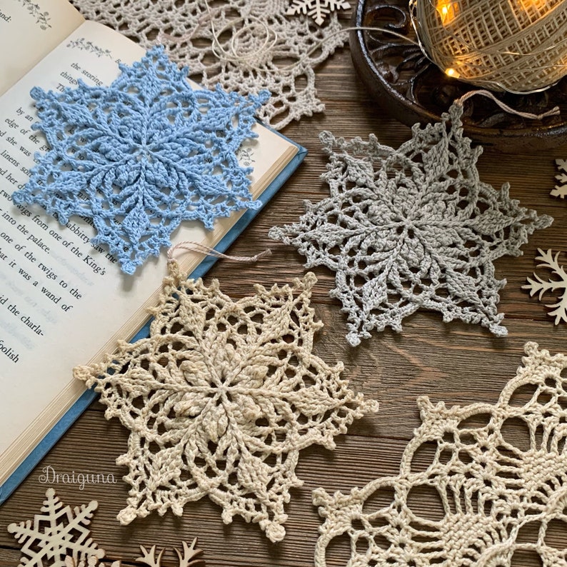 May include: Three crocheted snowflakes in different colors and patterns. The snowflakes are light blue, gray, and beige. They are arranged on a wooden surface with a book and other decorative items.