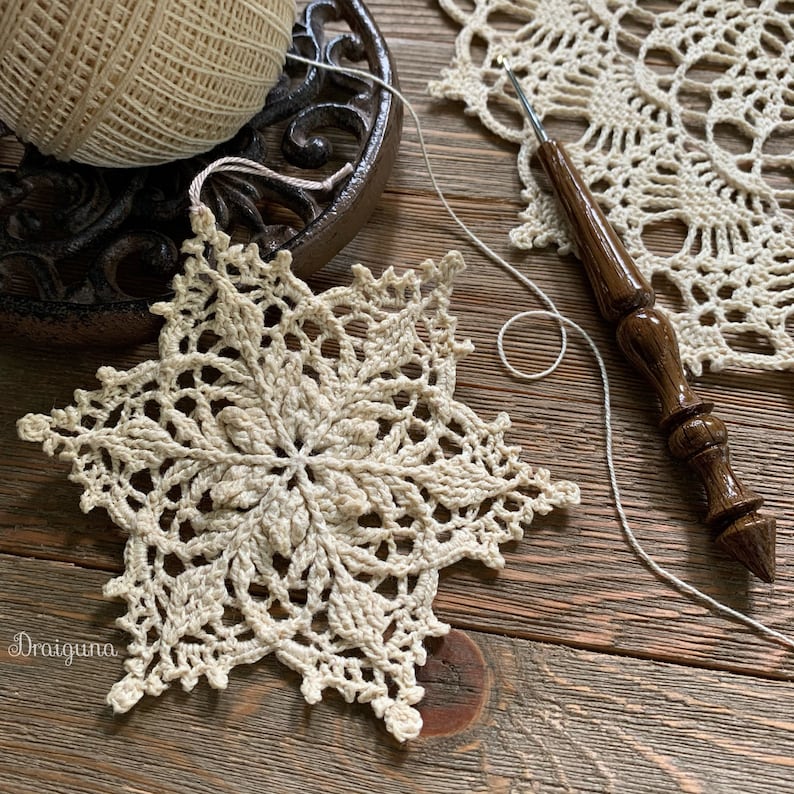 May include: A delicate crocheted snowflake ornament in a cream color, with a wooden crochet hook and a ball of yarn in the background.