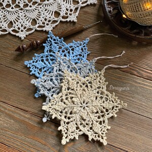 May include: Three crocheted snowflakes in shades of blue and beige. The snowflakes are layered on top of each other, with the largest snowflake on the bottom. The snowflakes are on a wooden surface.