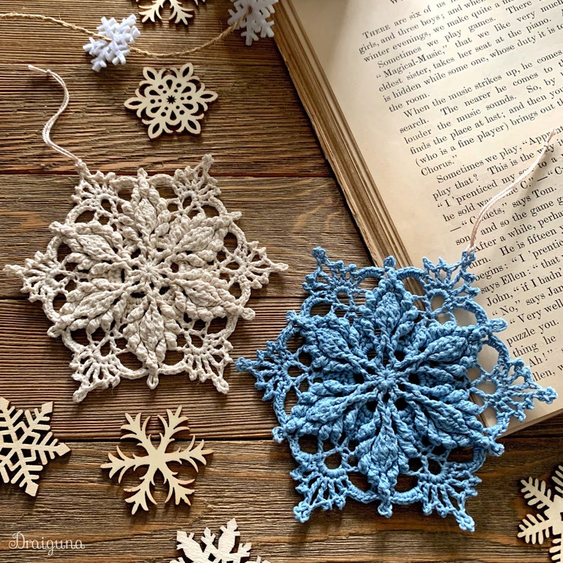 May include: Two crocheted snowflakes, one in beige and one in blue, are displayed on a wooden surface.  The snowflakes are surrounded by smaller wooden snowflakes.