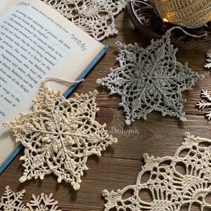 May include: Three crocheted snowflakes in white and gray, with a book and wooden snowflakes in the background. The snowflakes are delicate and intricate, with a variety of patterns and textures.