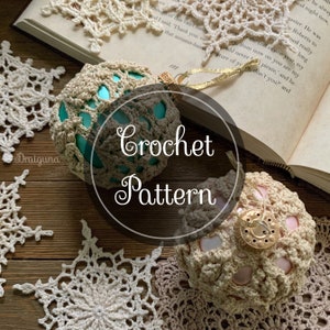 Puede incluir: Dos adornos de ganchillo, uno turquesa y otro rosa, con un copo de nieve de ganchillo blanco en primer plano. Los adornos están sobre una superficie de madera con un libro en el fondo. El texto "Crochet Pattern" está superpuesto a la imagen.