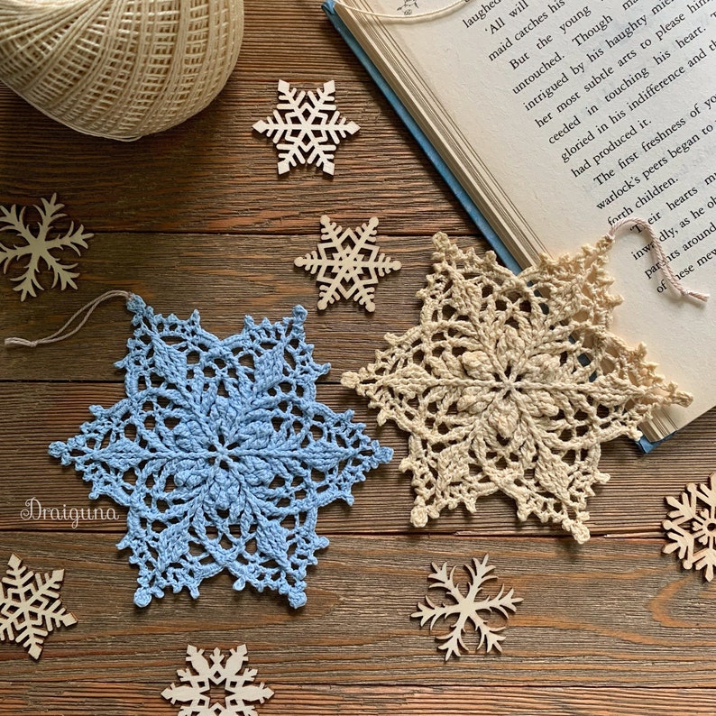May include: Two crocheted snowflakes, one blue and one beige, are displayed on a wooden surface with several wooden snowflake ornaments.