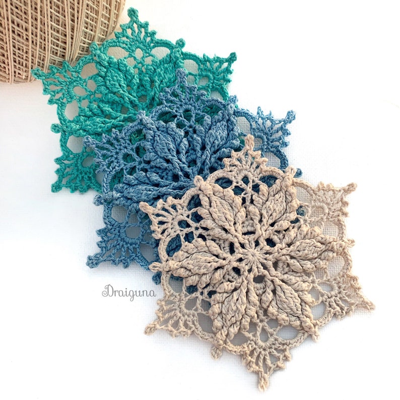 May include: Three crocheted snowflakes in different colors: teal, blue, and beige. The snowflakes are layered on top of each other, with the beige snowflake on top.