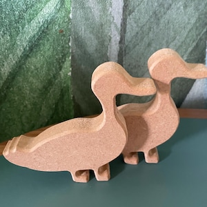 May include: Two wooden duck figurines. The ducks are light brown and have a simple, stylized design. They are standing on a green surface, with a green and white patterned background.