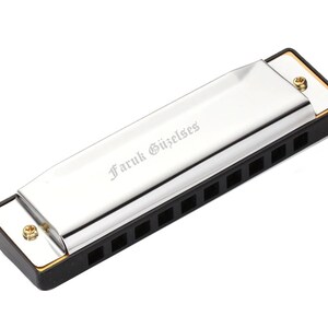 Pode incluir: Uma gaita de boca prateada com tampas de plástico preto e um bocal dourado. A gaita de boca tem o nome "Faruk Güzelses" gravado na parte superior.