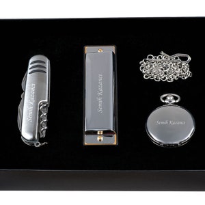 K&ouml;nnte beinhalten: Eine schwarze Geschenkbox mit einem silbernen Taschenmesser, einer Mundharmonika und einer Taschenuhr. Jedes Element ist mit dem Namen "Semih Kazanca" graviert.
