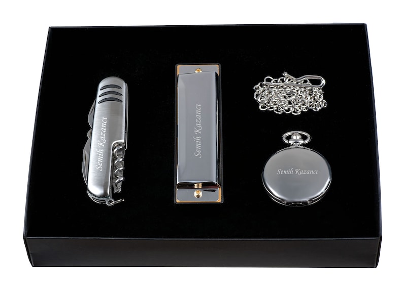 K&ouml;nnte beinhalten: Eine schwarze Geschenkbox mit einem silbernen Taschenmesser, einer Mundharmonika und einer Taschenuhr. Jedes Element ist mit dem Namen "Semih Kazanca" graviert.