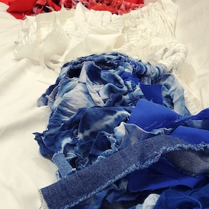 Pu&ograve; includere: Una composizione astratta di ritagli di tessuto nei toni del rosso, bianco e blu. La disposizione comprende varie trame, come denim, tie-dye e tessuti semplici, creando un effetto visivo stratificato e strutturato. I tessuti sono disposti su una superficie bianca.
