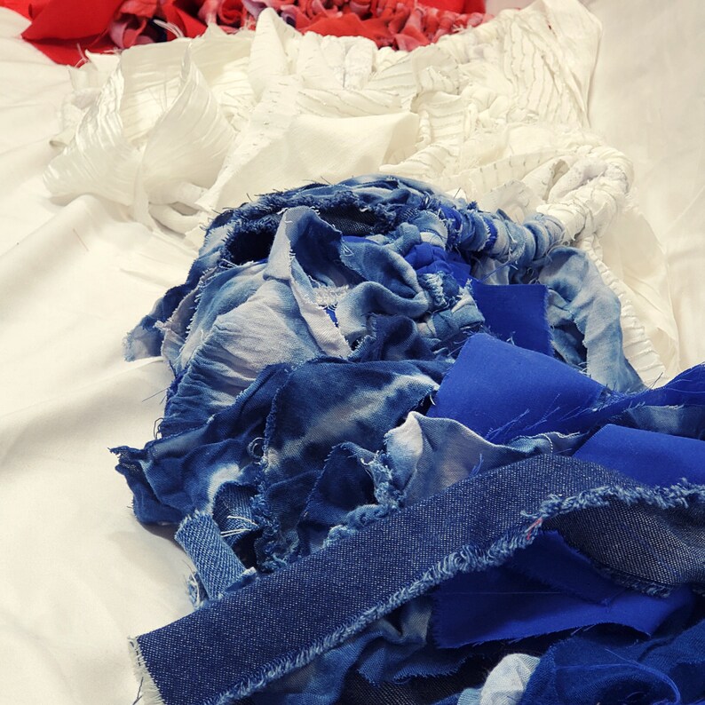 Pu&ograve; includere: Una composizione astratta di ritagli di tessuto nei toni del rosso, bianco e blu. La disposizione comprende varie trame, come denim, tie-dye e tessuti semplici, creando un effetto visivo stratificato e strutturato. I tessuti sono disposti su una superficie bianca.