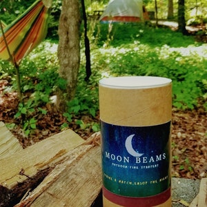 Op de afbeelding: Een cilindervormige kartonnen koker met een etiket met een halve maan en een sterrenhemel. De tekst luidt MOON BEAMS OUTDOOR FIRE STARTERS en STRIKE A MATCH, ENJOY THE NIGHT. De koker heeft een witte deksel en een donkerrode band aan de onderkant.