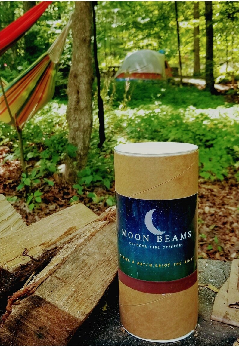 Op de afbeelding: Een cilindervormige kartonnen koker met een etiket met een halve maan en een sterrenhemel. De tekst luidt MOON BEAMS OUTDOOR FIRE STARTERS en STRIKE A MATCH, ENJOY THE NIGHT. De koker heeft een witte deksel en een donkerrode band aan de onderkant.