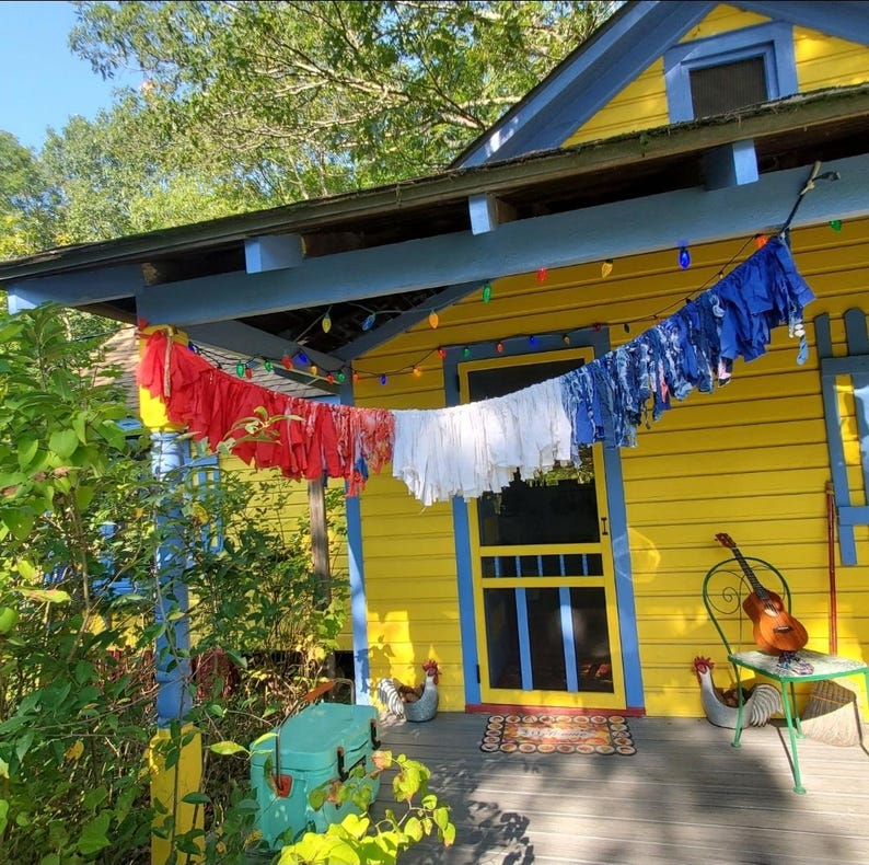 Pu&ograve; includere: Una casa gialla con finiture blu e un portico decorato con biancheria colorata appesa a un filo. Il portico ha una ghiacciaia verde, un tavolino con una chitarra e galline decorative.