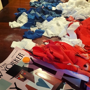 Pu&ograve; includere: Ritagli di tessuto rosso, bianco e blu sparsi su una superficie di legno. La copertina della rivista "The New Yorker", datata 31 maggio 2021, &egrave; visibile, con un'illustrazione colorata.