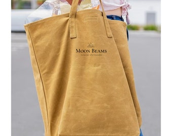 Può includere: Una borsa tote in tela color senape con manici robusti. La borsa presenta il testo "MOON BEAMS LUXURY OUTDOORS" in nero. La borsa è piena di oggetti avvolti in plastica trasparente. La borsa è portata da una persona che indossa jeans blu.