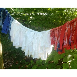 Pu&ograve; includere: Una ghirlanda festosa fatta di strisce di tessuto rosse, bianche e blu, appesa su uno sfondo di fogliame verde. Le strisce sono di diverse trame e lunghezze, creando un effetto decorativo strutturato.