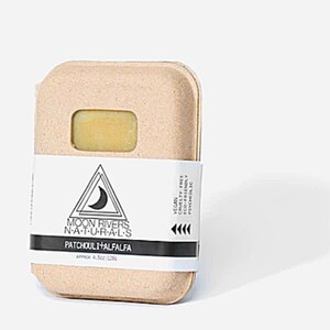 May include: A bar of soap in a cardboard container. The soap is light yellow and visible through a rectangular window. The label reads "MOON RIVERS NATURALS PATCHOULI + ALFALFA" and "APPROX 4.5oz | 128g". The label also states "VEGAN CRUELTY FREE ECO-FRIENDLY PSYCHEDLIC".