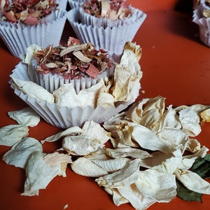 Op de afbeelding: Een wit papieren cupcakevormpje gevuld met een mengsel van gedroogde rode en bruine houtsnippers, omringd door verspreide gedroogde witte rozenblaadjes. Andere gevulde vormpjes zijn zichtbaar op de achtergrond op een oranje ondergrond.