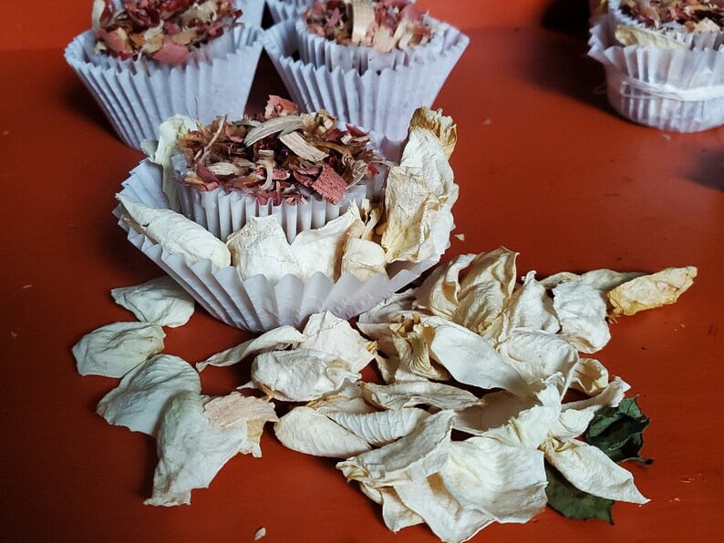 Op de afbeelding: Een wit papieren cupcakevormpje gevuld met een mengsel van gedroogde rode en bruine houtsnippers, omringd door verspreide gedroogde witte rozenblaadjes. Andere gevulde vormpjes zijn zichtbaar op de achtergrond op een oranje ondergrond.