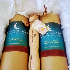 Op de afbeelding: Twee cilindrische kartonnen kokers met het label Moon Beams Outdoor Fire Starters met een halve maan afbeelding en een houten handvat met een metalen aansteker bevestigd aan een leren koord, in wit vloeipapier in een doos.