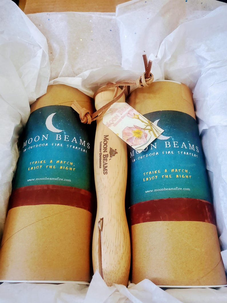 Op de afbeelding: Twee cilindrische kartonnen kokers met het label Moon Beams Outdoor Fire Starters met een halve maan afbeelding en een houten handvat met een metalen aansteker bevestigd aan een leren koord, in wit vloeipapier in een doos.