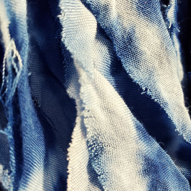 Pu&ograve; includere: Primo piano di un tessuto blu e bianco con una trama strutturata. Il tessuto ha un aspetto invecchiato con bordi sfilacciati, creando un'estetica vintage o boh&eacute;mien. La sfumatura di colore passa dal blu intenso a tonalit&agrave; pi&ugrave; chiare.