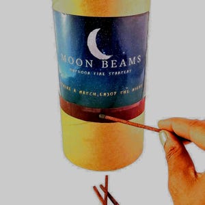 Op de afbeelding: Een cilindervormige kartonnen aanmaakblokje met een gele buitenkant en een centraal label met een maansikkel, een sterrenhemel en de tekst MOON BEAMS OUTDOOR FIRE STARTERS STRIKE A MATCH, ENJOY THE NIGHT, inclusief drie losse lucifers.