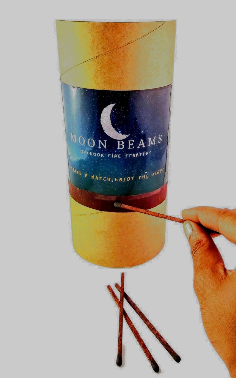 Op de afbeelding: Een cilindervormige kartonnen aanmaakblokje met een gele buitenkant en een centraal label met een maansikkel, een sterrenhemel en de tekst MOON BEAMS OUTDOOR FIRE STARTERS STRIKE A MATCH, ENJOY THE NIGHT, inclusief drie losse lucifers.