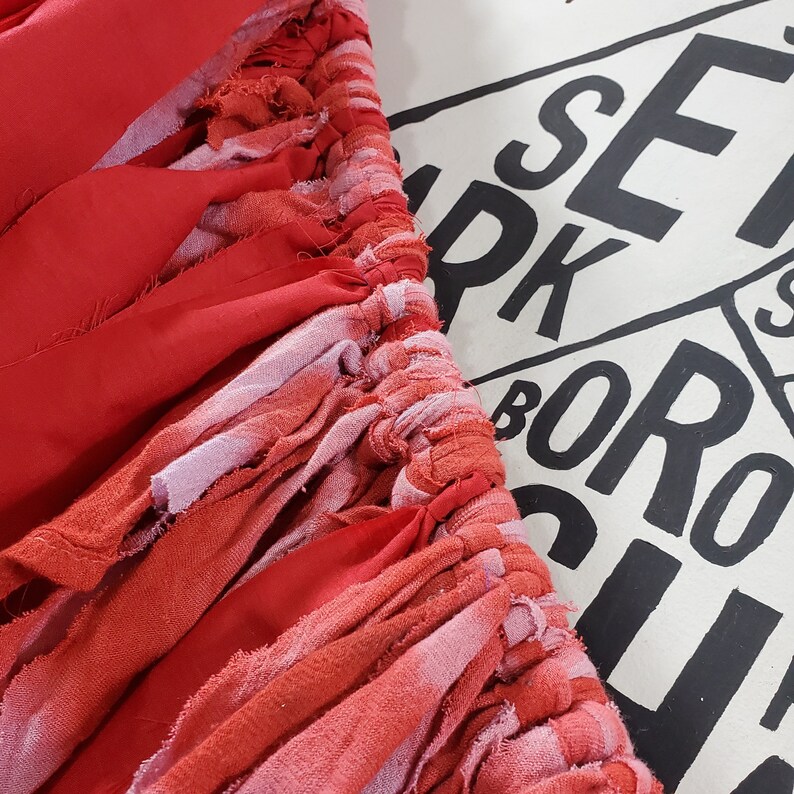 Pu&ograve; includere: Primo piano di un tessuto rosso con un design a strati. Il tessuto &egrave; composto da strisce di varie tonalit&agrave; di rosso e rosa, con un bordo strutturato e sfilacciato. Lo sfondo presenta testo nero su una superficie bianca.