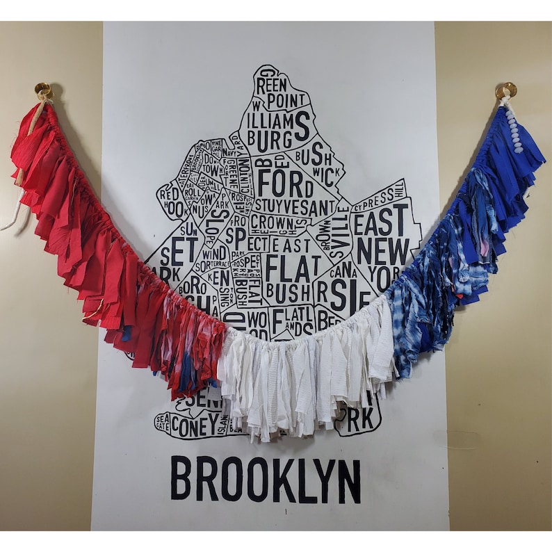 Pu&ograve; includere: Una ghirlanda a tema patriottico con strisce di tessuto rosse, bianche e blu. La ghirlanda &egrave; appesa davanti a una mappa in bianco e nero di Brooklyn, con la parola "BROOKLYN" in basso.