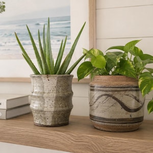 May include: Two ceramic planters containing live plants. The left planter is tall, cylindrical, and speckled grey with a ribbed texture. The right planter is shorter, rounded, and features horizontal brown wavy lines on a light grey base.