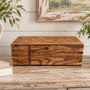 May include: A rectangular wooden box with a visible natural grain pattern and a dark brown finish. The box features a hinged lid with a small, centered dark metal knob for opening. The wood displays a textured surface with variations in color.