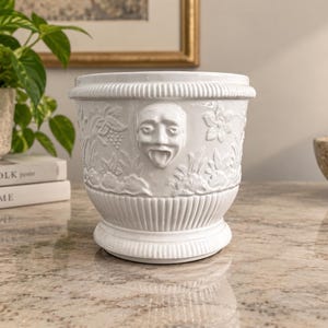 May include: A white ceramic planter featuring a relief design of a face with an open mouth and tongue extended, surrounded by floral and fruit motifs. The vessel has a fluted base and a decorative rim with vertical lines around the top edge.