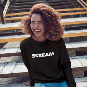 May include: A woman wearing a black long-sleeved sweatshirt with the word "SCREAM" printed in white on the front. She is smiling and sitting on a set of stairs.
