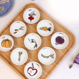 May include: A set of nine small white ceramic bowls arranged on a square bamboo tray. Each bowl features a unique hand-drawn illustration of a food item, including a beet, carrot, pumpkin, fish, pomegranate, leek, beans, and apple, with Hebrew text.