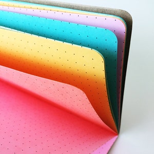 Puede incluir: Un cuaderno multicolor con páginas de puntos. Las páginas son rosa, naranja, amarillo, turquesa y lavanda. El cuaderno está abierto y las páginas están desplegadas.