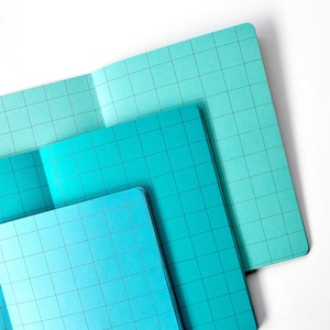 May include: Three blue grid notebooks stacked on top of each other. The top notebook is a lighter shade of blue than the bottom two notebooks.