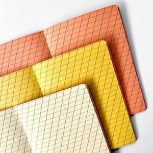 May include: Three notebooks with grid patterns. The top notebook is orange, the middle notebook is yellow, and the bottom notebook is white.