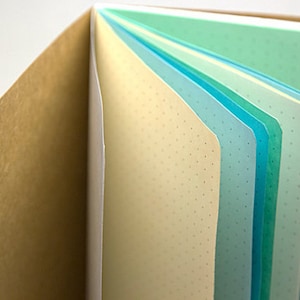 May include: A brown notebook with a textured cover is open to reveal a stack of colorful pages with dotted lines. The pages are in shades of green, blue, and white.