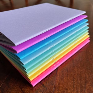 Puede incluir: Una pila de cuadernos coloridos con cubiertas en tonos morado, rosa, azul, verde, amarillo y rosa. Cada cuaderno está encuadernado con grapas a lo largo del lomo. Los cuadernos están apilados sobre una superficie de madera.