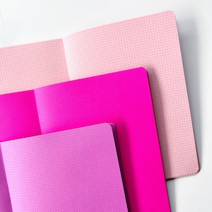 Könnte beinhalten: Drei Notizbücher mit gepunkteten Seiten. Das obere Notizbuch ist rosa, das mittlere Notizbuch ist magenta und das untere Notizbuch ist ein Farbverlauf von rosa und lila.