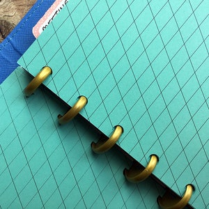 May include: A turquoise blue notebook with a grid pattern and gold rings. The notebook is open and the rings are visible.