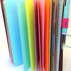May include: A brown leather bound notebook with colorful pages. The pages are in a rainbow pattern, with blue, green, yellow, pink, orange, and red pages. The notebook is open and the pages are fanned out.