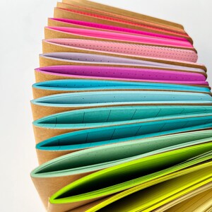 May include: A stack of colorful notebooks with different patterned paper inside. The notebooks have brown covers and are arranged in a rainbow pattern.