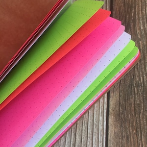 May include: A close-up of a notebook with multiple colored pages. The pages are pink, green, white, and red. The pages are dotted with small black dots.