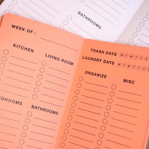 May include: A pink and white checklist notepad with sections for kitchen, living room, bedrooms, bathrooms, trash days, laundry days, organise, and misc. The notepad has a week of section at the top.