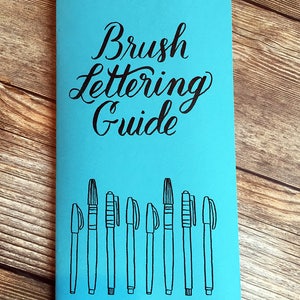Op de afbeelding: Een blauw notitieboekje met de tekst "Brush Lettering Guide" in zwarte cursieve letters. Het notitieboekje heeft een rij van tien zwarte penseelpennen die op de voorkant zijn getekend.